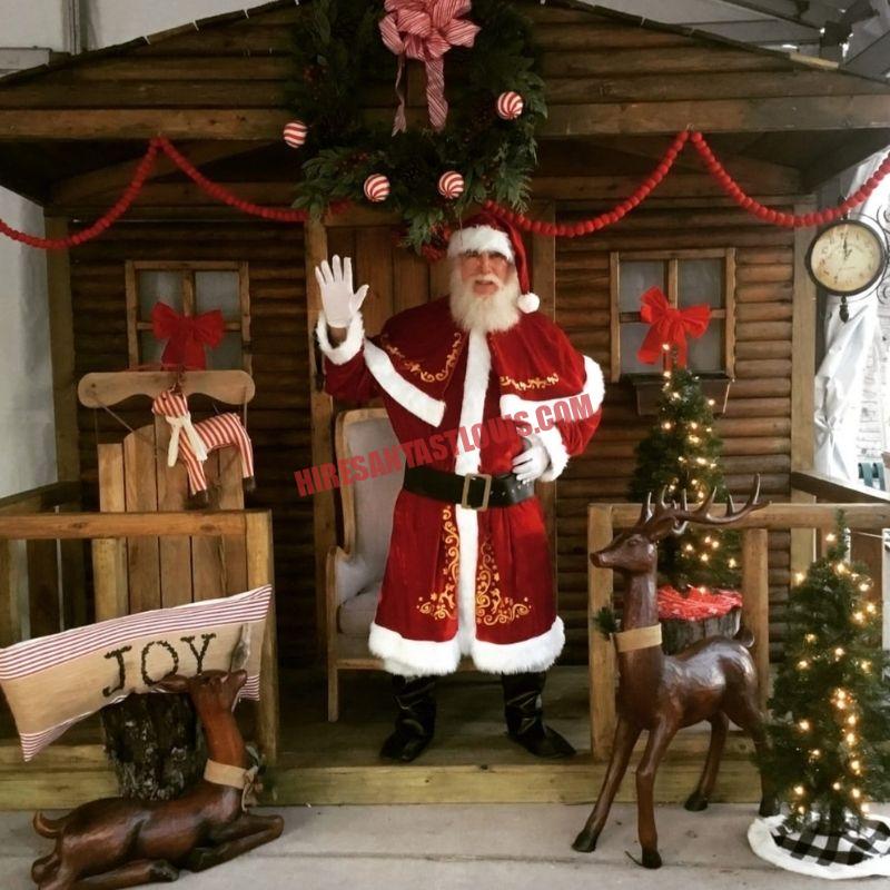 Real beard Santa for hire in St. Louis waving from a log-cabin doorway with wooden reindeer.