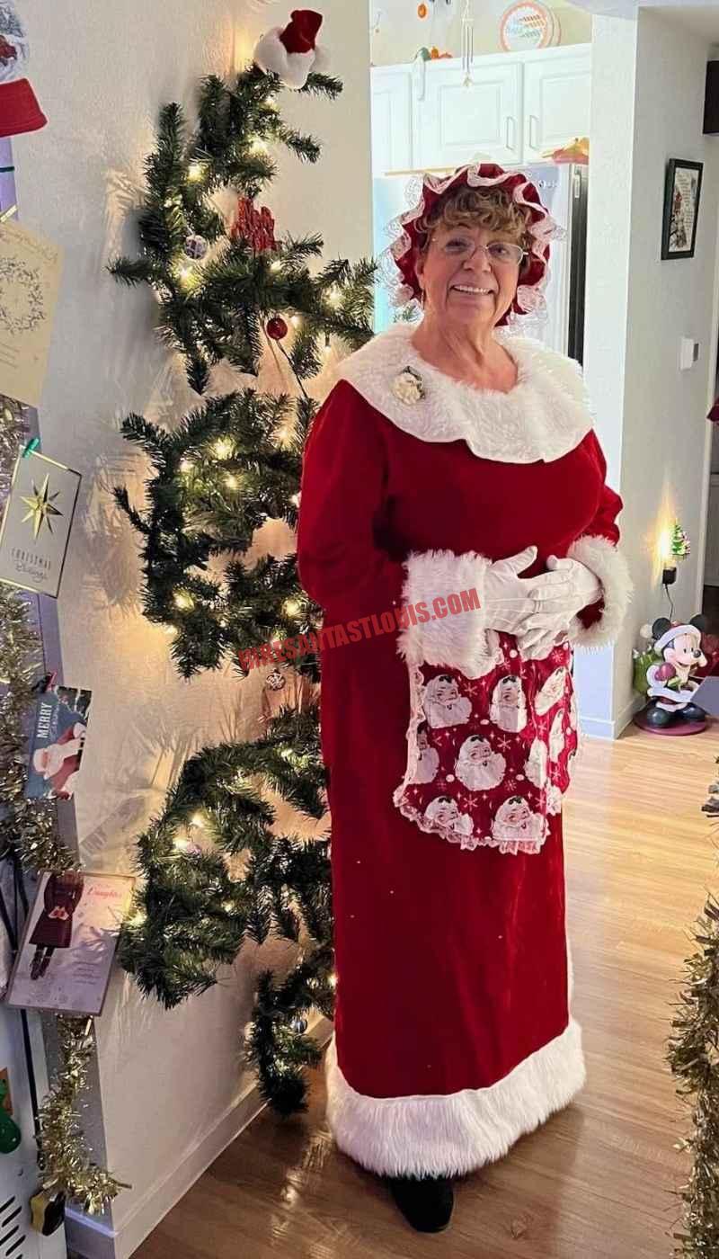 Mrs. Claus for hire in St. Louis standing in a hallway by a slim lit tree in a red dress.
