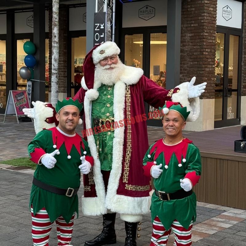 Santa Claus impersonator for hire in St. Louis standing outdoors with two elves, thumbs up.
